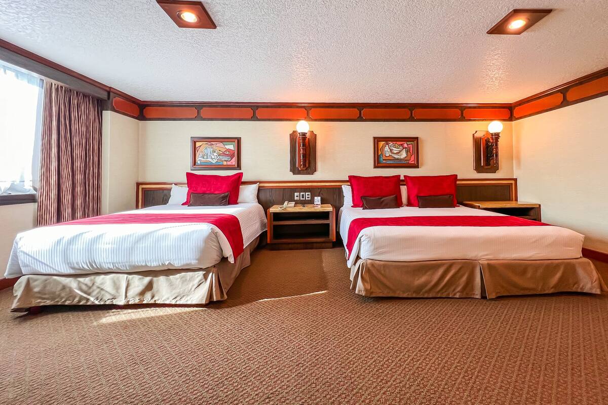 A hotel room with two beds, each with red and white bedding, wooden headboards, and wall-mounted lamps.