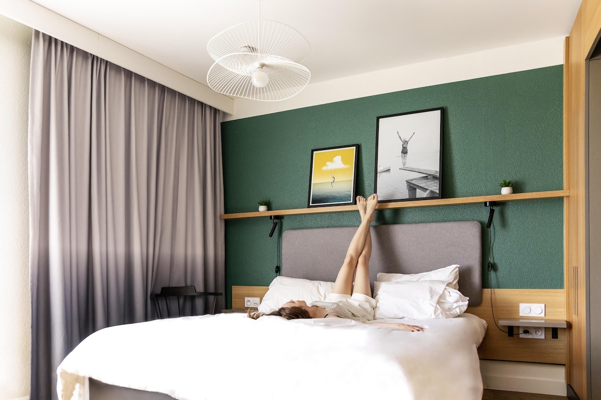 A bed with a person lying on it, a ceiling fan, a wall with two framed pictures, and a window with curtains.