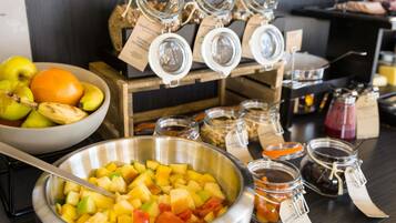 A buffet setup with a bowl of fruit salad, jars of preserves, and a fruit bowl with apples and oranges.