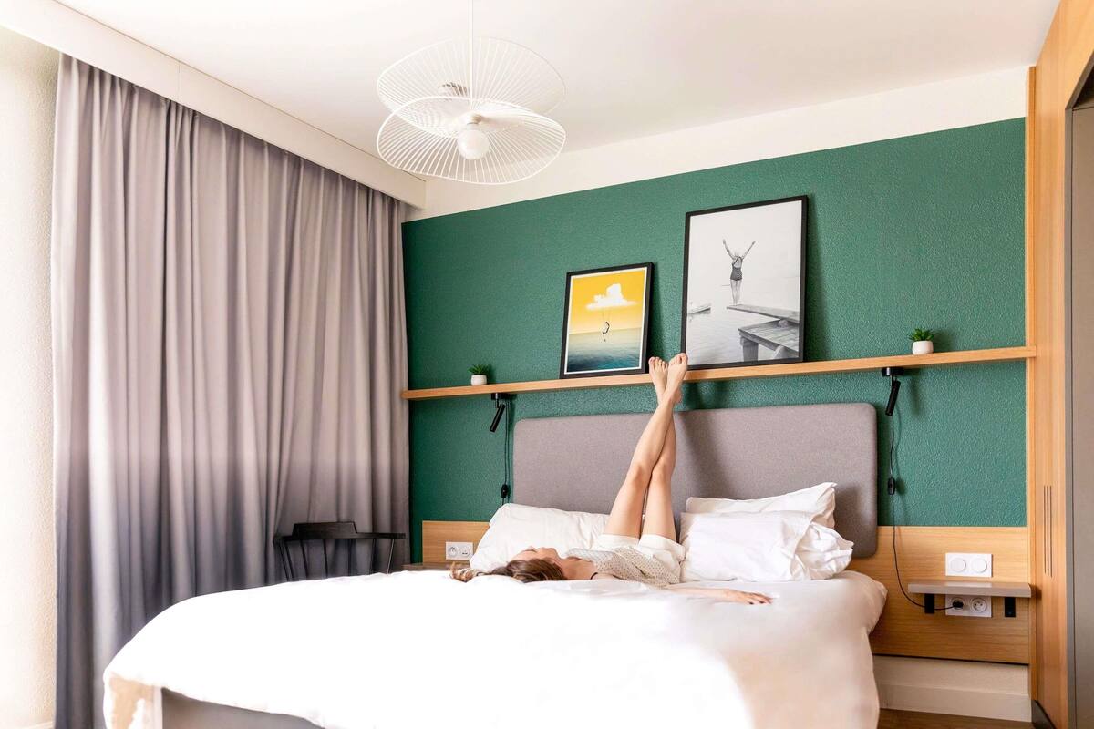 Een moderne hotelkamer met een bed, hoofdbord, een wandplank met kunstwerk, een plafondventilator en een raam met gordijnen.