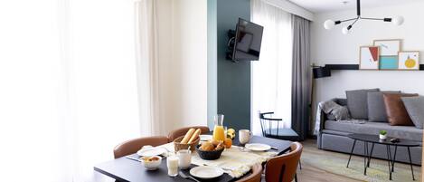A modern living room with a dining area, featuring a flat-screen TV, a sofa with cushions, a coffee table, and a dining table set with breakfast items.