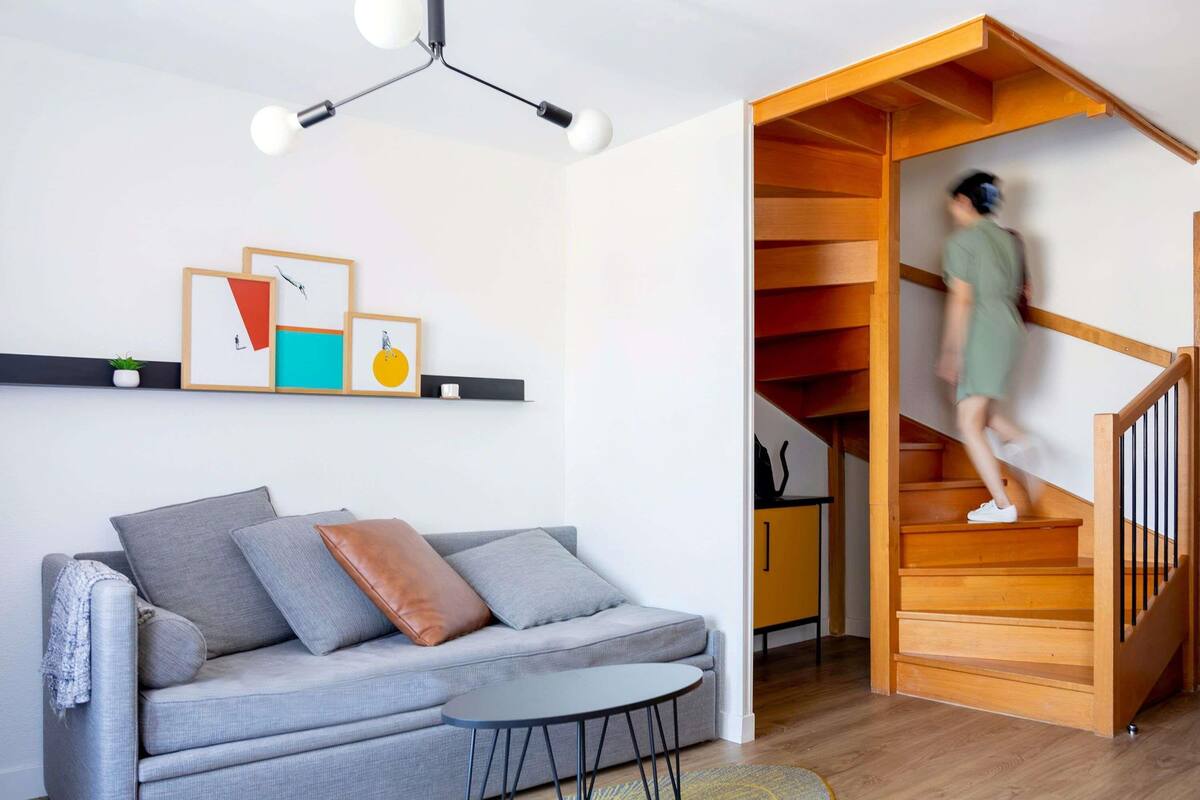 Een moderne woonkamer met een grijze bank, een klein tafeltje, een trap die naar een lager gelegen niveau leidt, en een plafondlamp.
