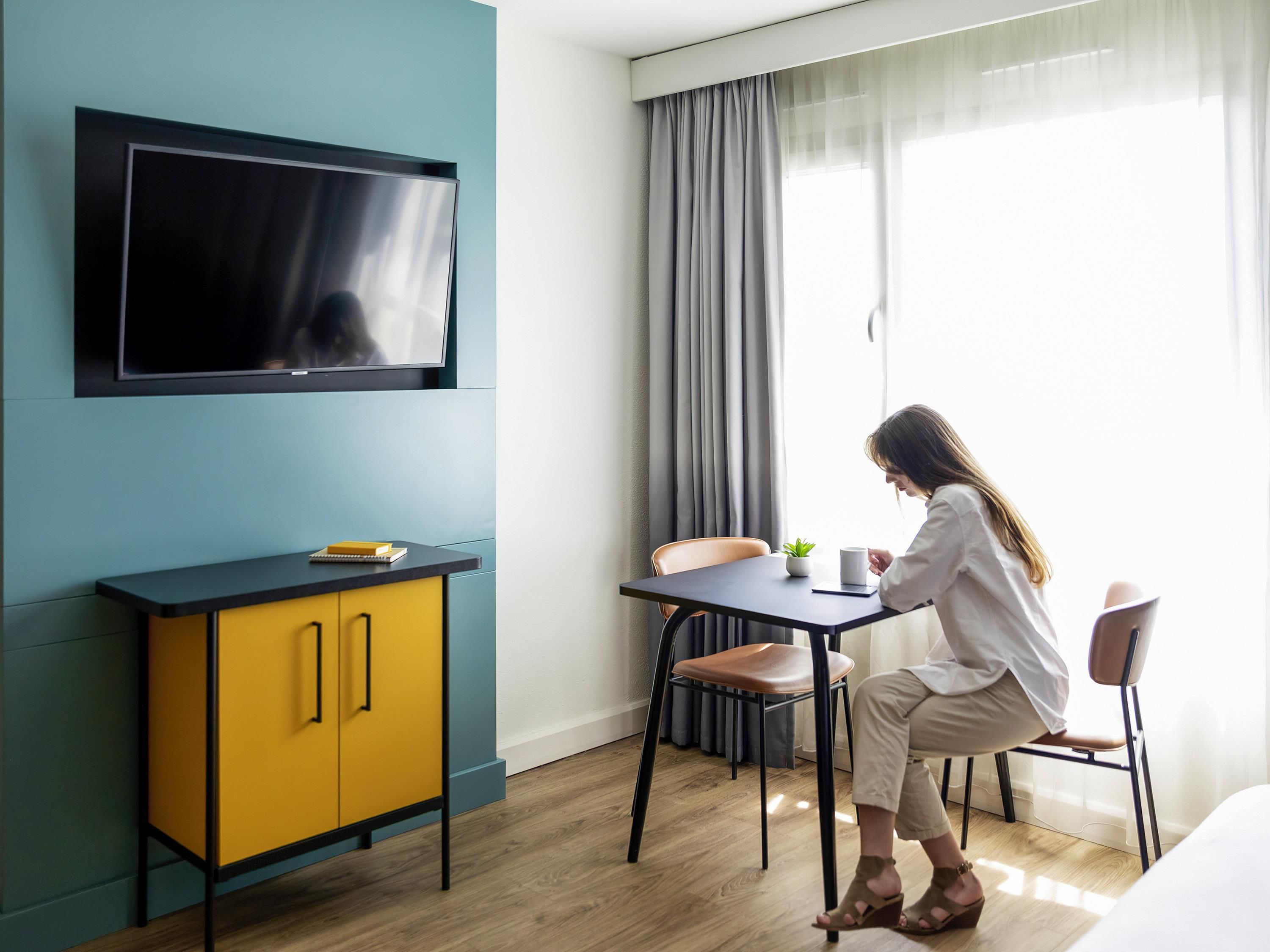 Une chambre d’hôtel moderne avec un téléviseur fixé au mur, une petite table agrémentée d’une plante et une fenêtre avec des rideaux.