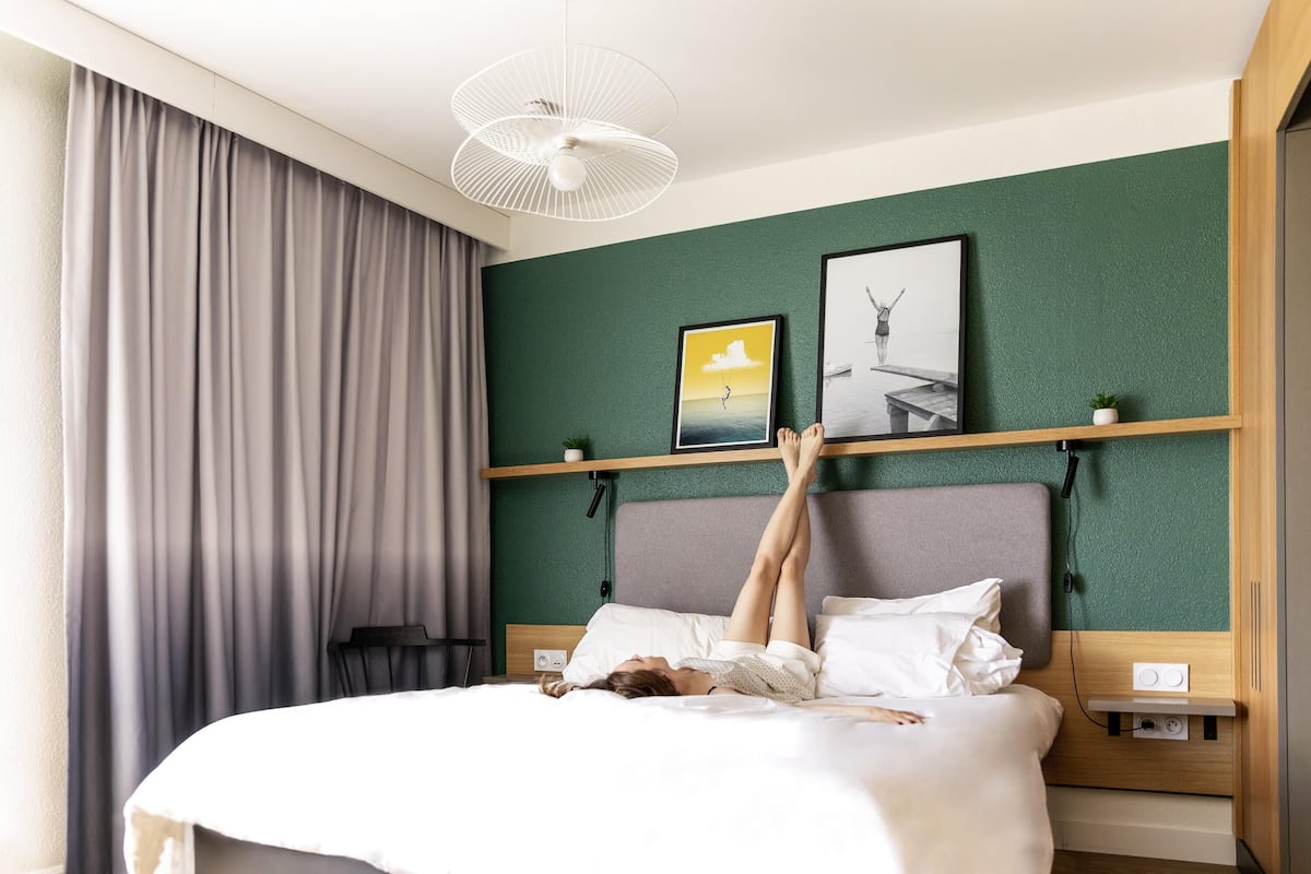 A bed with a person lying on it, a ceiling fan, a wall with two framed pictures, and a window with curtains.