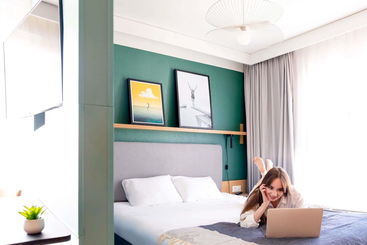 Een hotelkamer met een bed, een bureau met een laptop, een potplant en kunst aan de muur.
