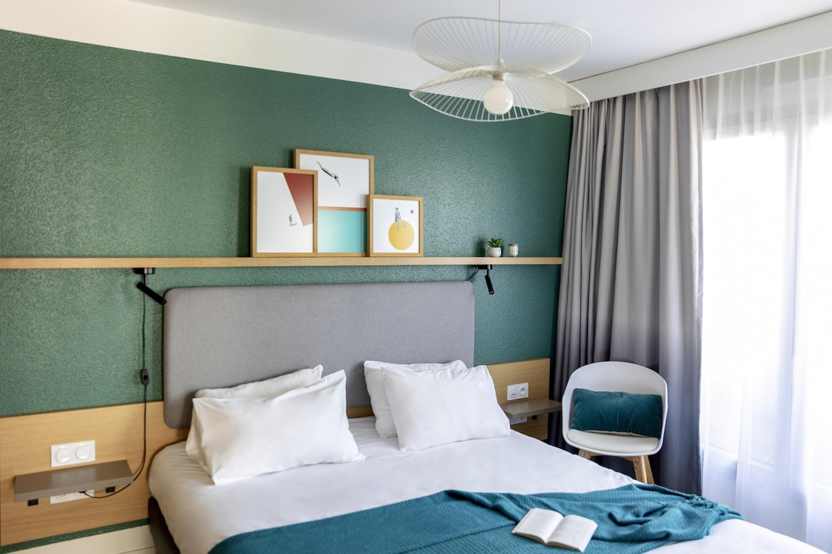 A hotel room with a bed, a bedside table, a chair, a ceiling fan, and artwork on the wall.