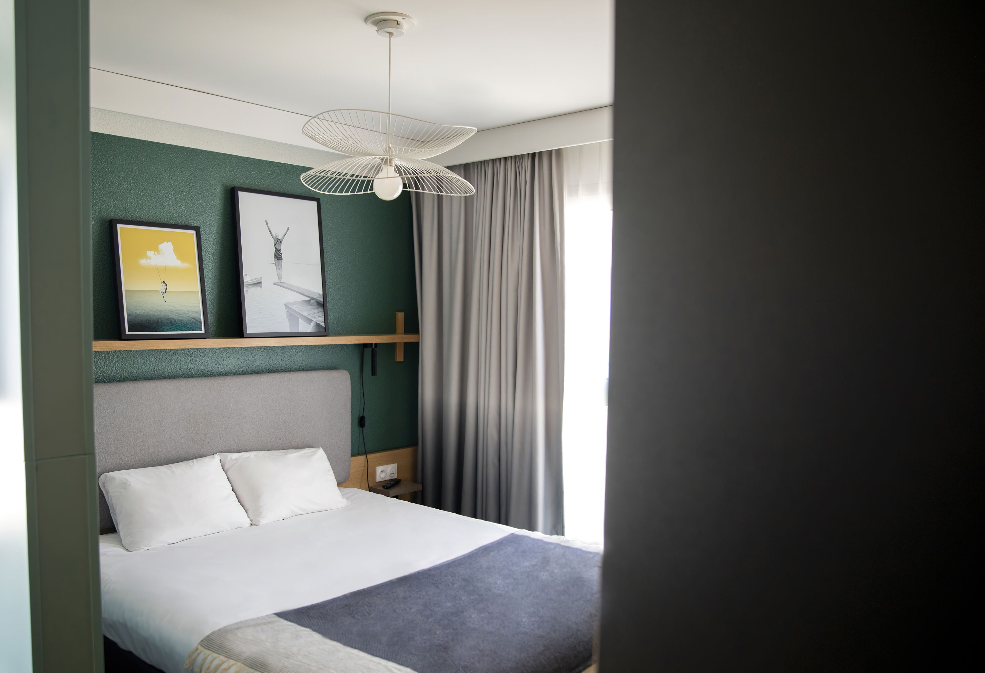 Quarto de hotel bem arrumado com cama, cabeceira, prateleira na parede e janela com cortinas.