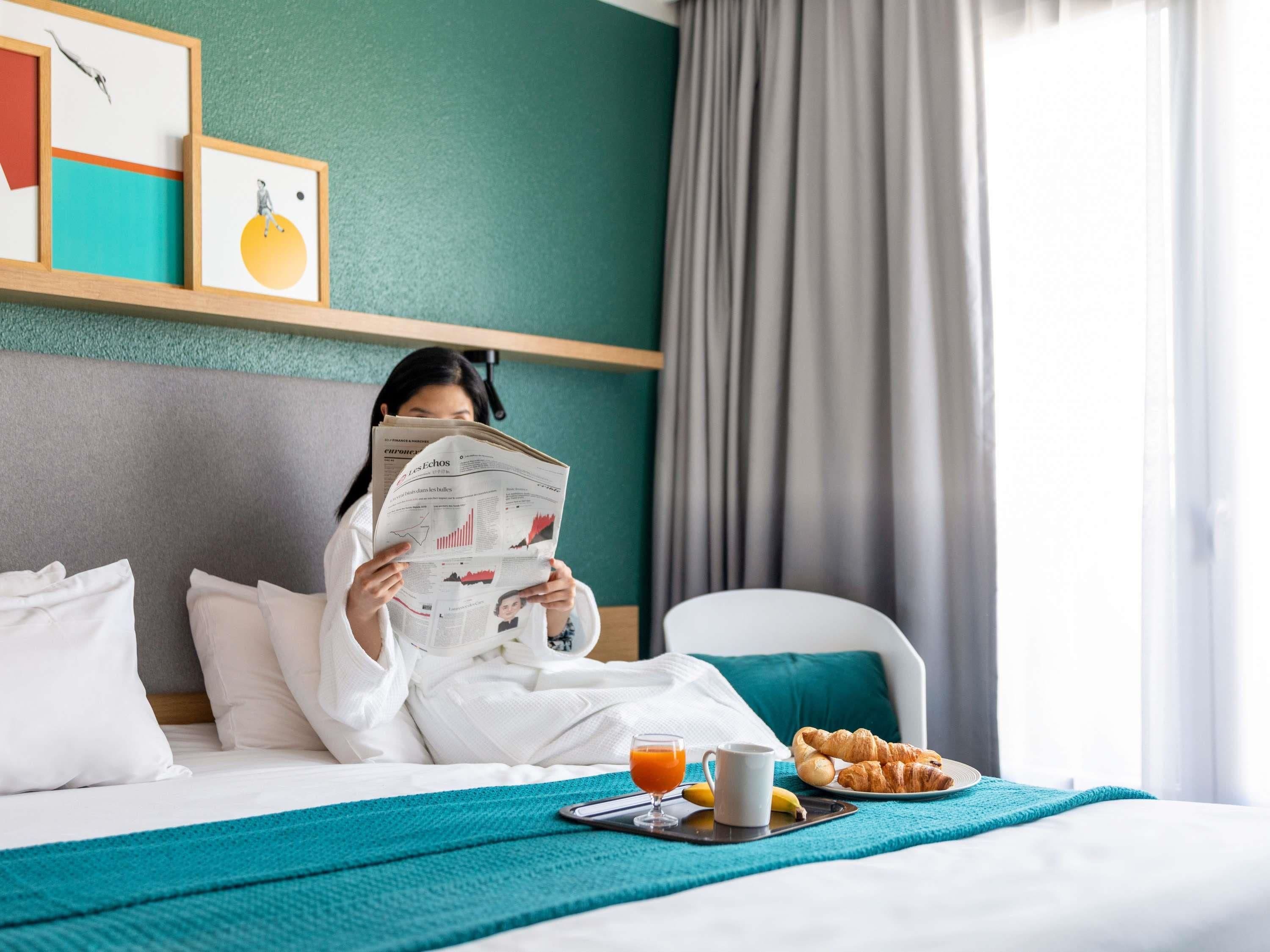 A hotel room with a bed, a breakfast tray with croissants, a newspaper, and a glass of orange juice.