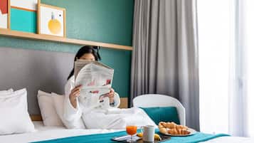 A hotel room with a bed, a breakfast tray with croissants, a newspaper, and a glass of orange juice.