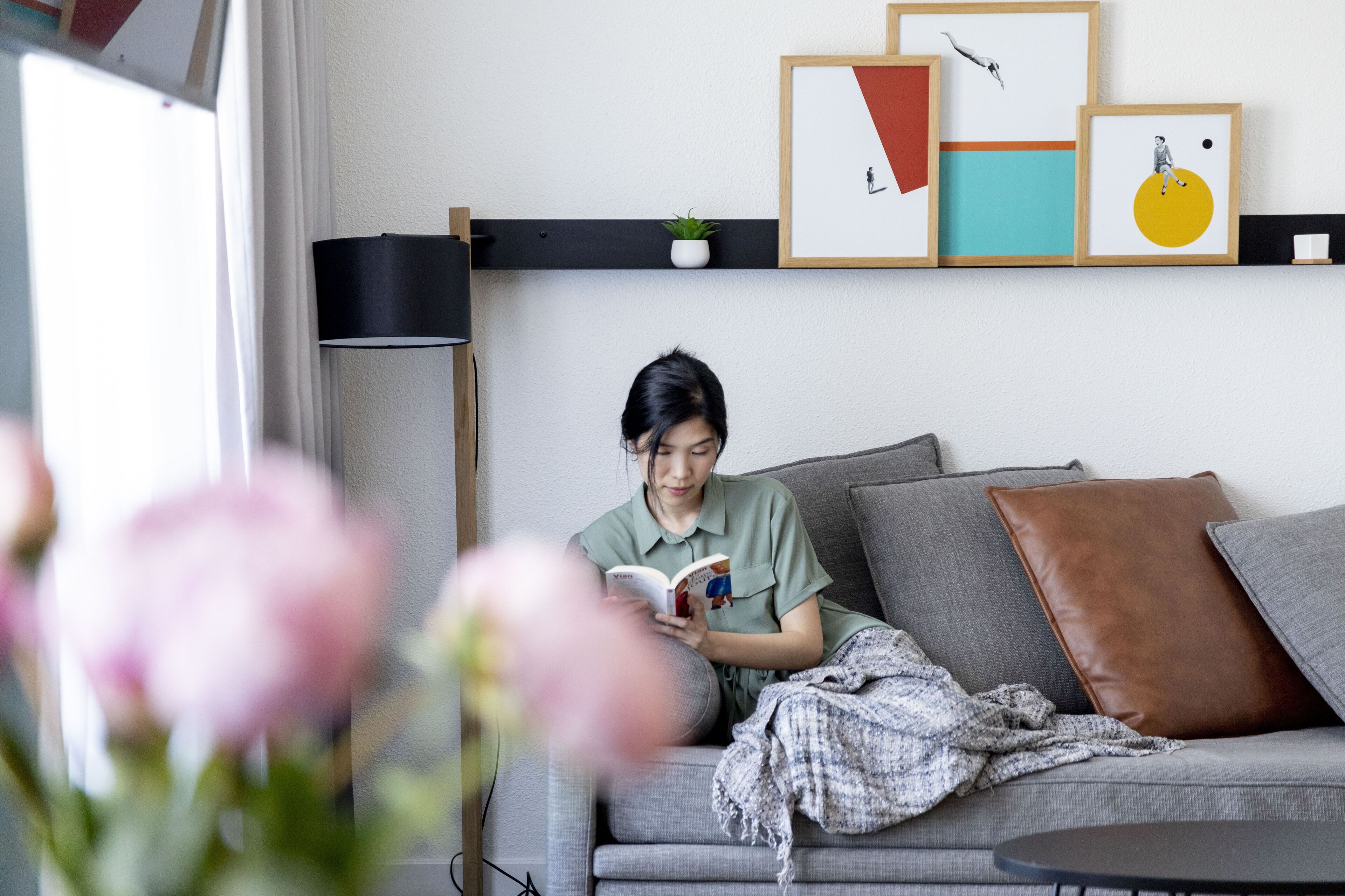 A person sitting on a grey sofa reading a book, with a black floor lamp and framed artwork on the wall.