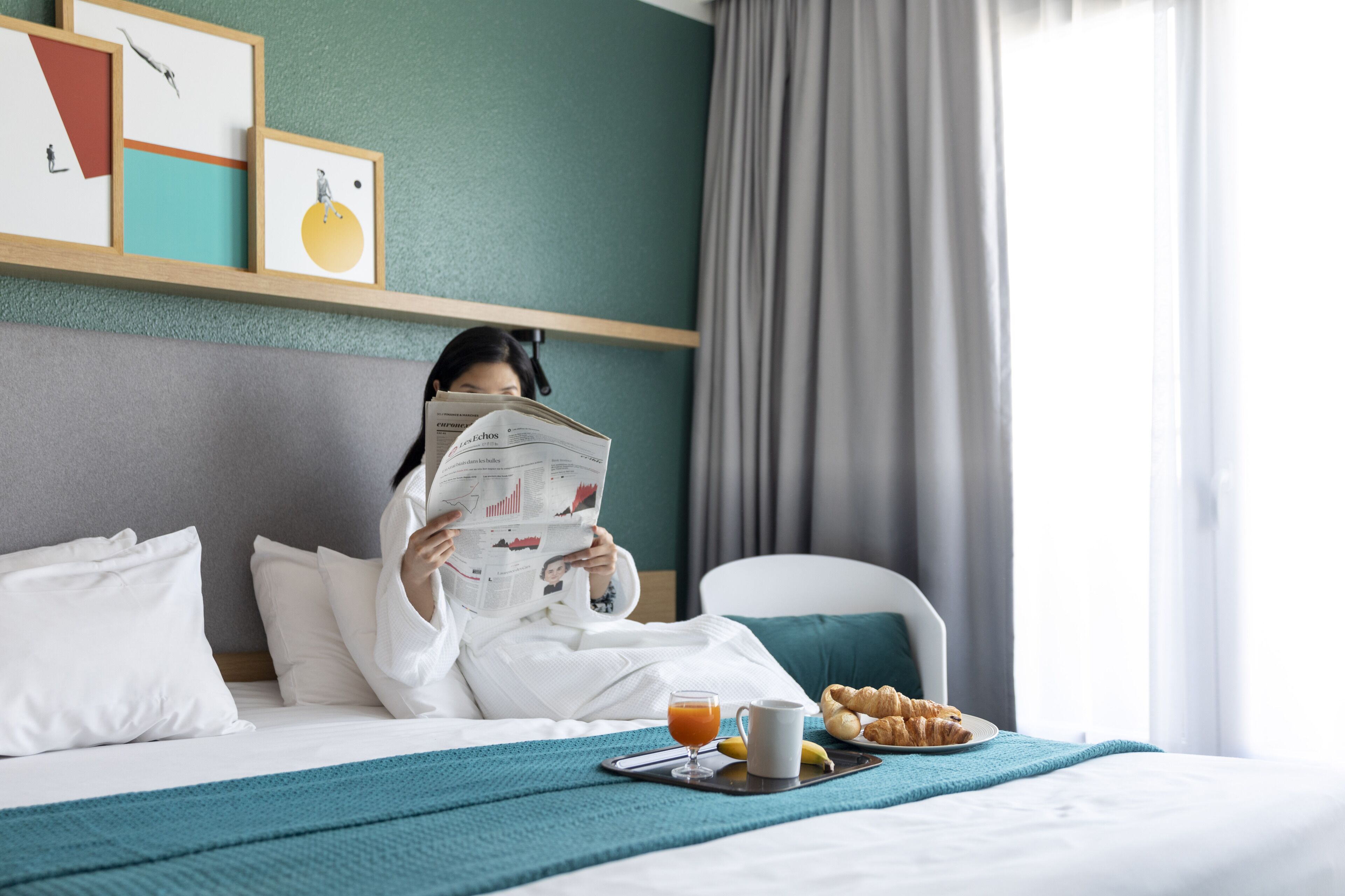 Ett hotellrum med en säng, ett nattduksbord med ett bricka med frukost, en tidning och konstverk på väggen.
