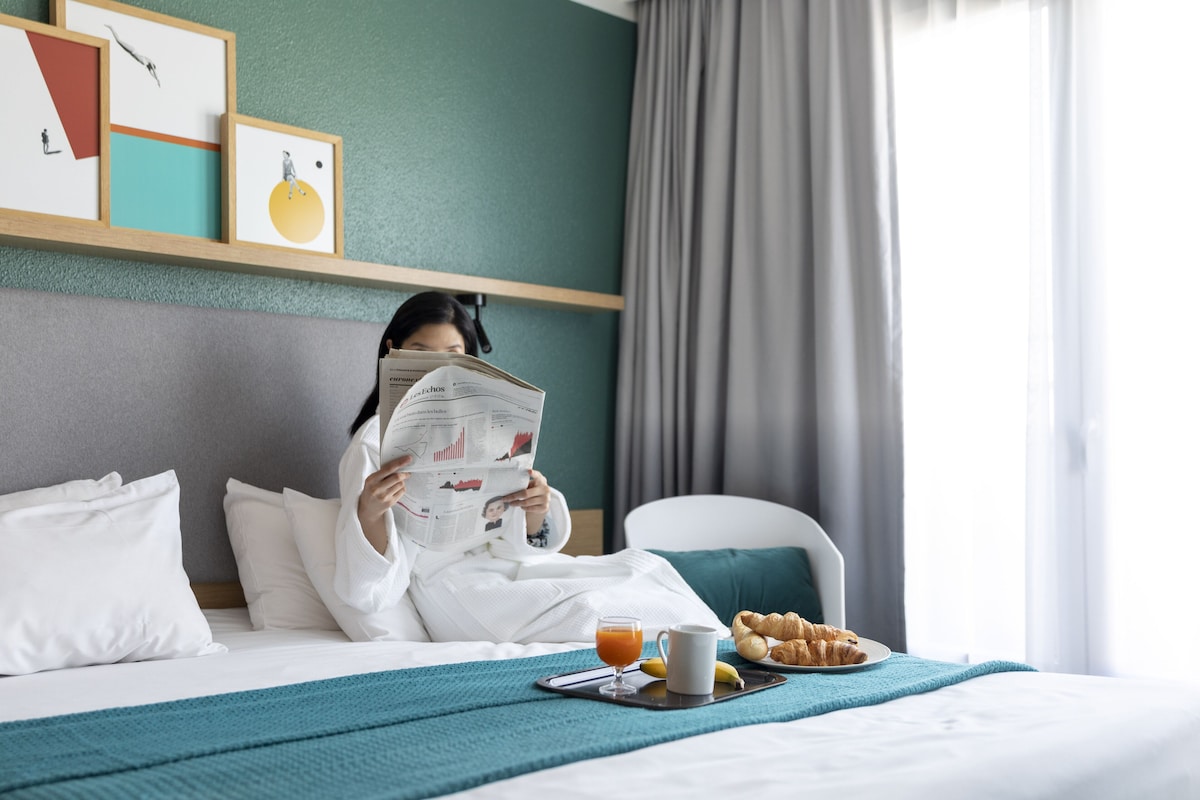 Habitación de hotel con cama, mesita de noche con bandeja de desayuno, un periódico y una obra de arte en la pared.