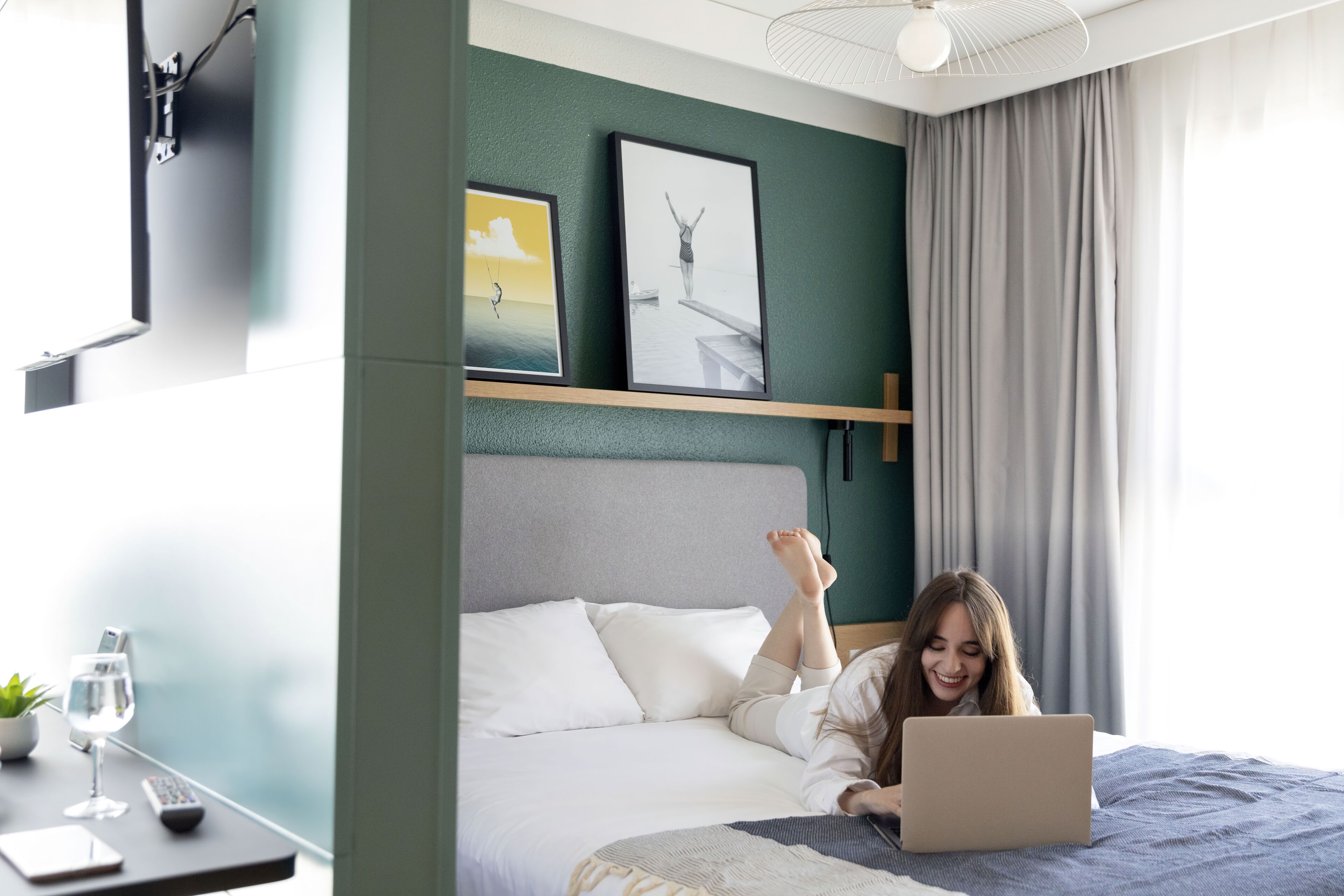 Un dormitorio con una cama, una laptop, un control remoto, un vaso de agua y una planta en una mesita de noche.
