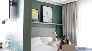 A bedroom with a bed, a laptop, a remote, a glass of water, and a plant on a bedside table.