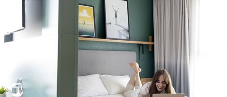 A bedroom with a bed, a laptop, a remote, a glass of water, and a plant on a bedside table.
