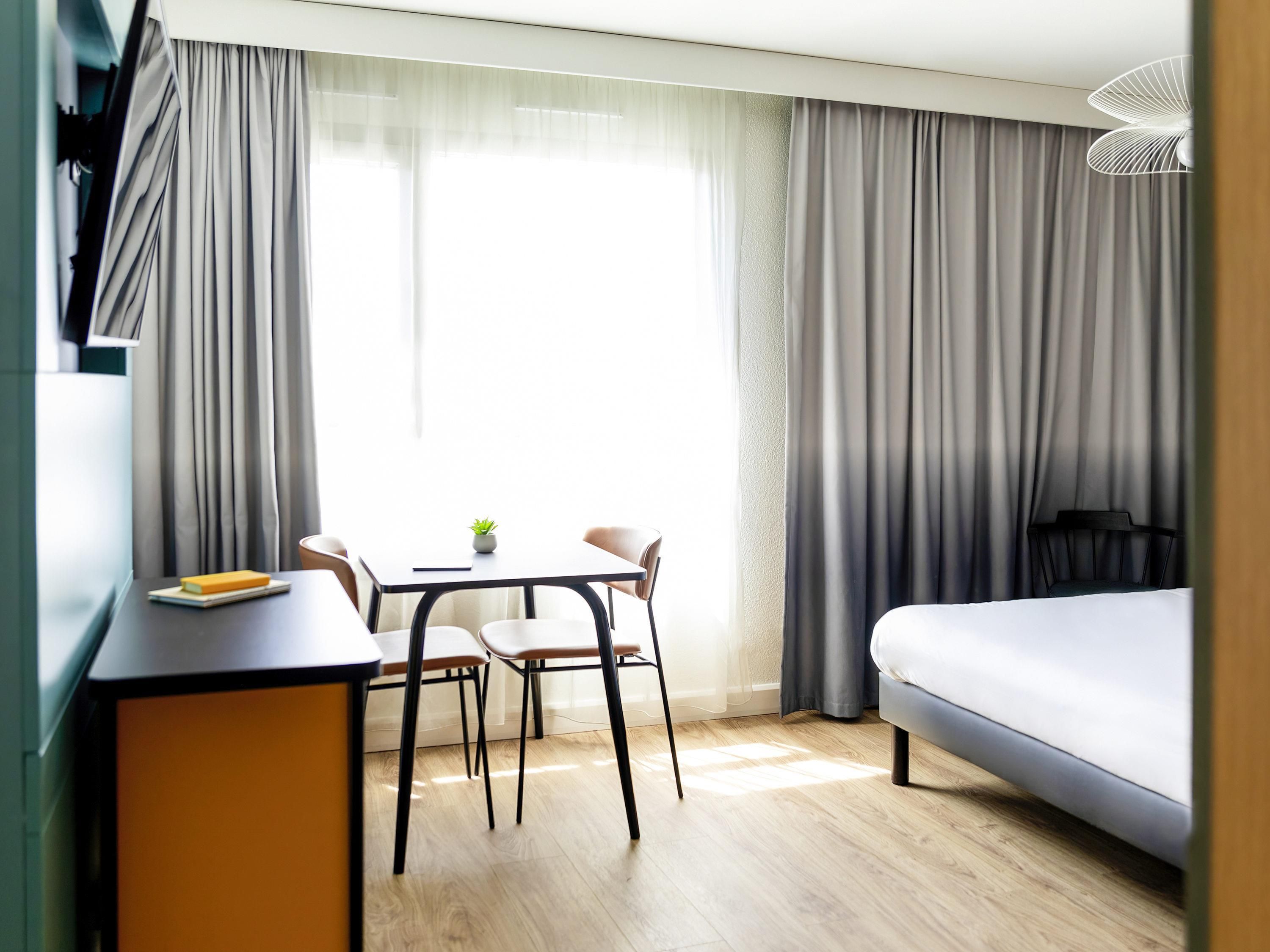 Une chambre d’hôtel moderne, équipée d’un lit, d’un bureau avec une chaise, d’une petite table avec une plante et d’une fenêtre avec des rideaux.