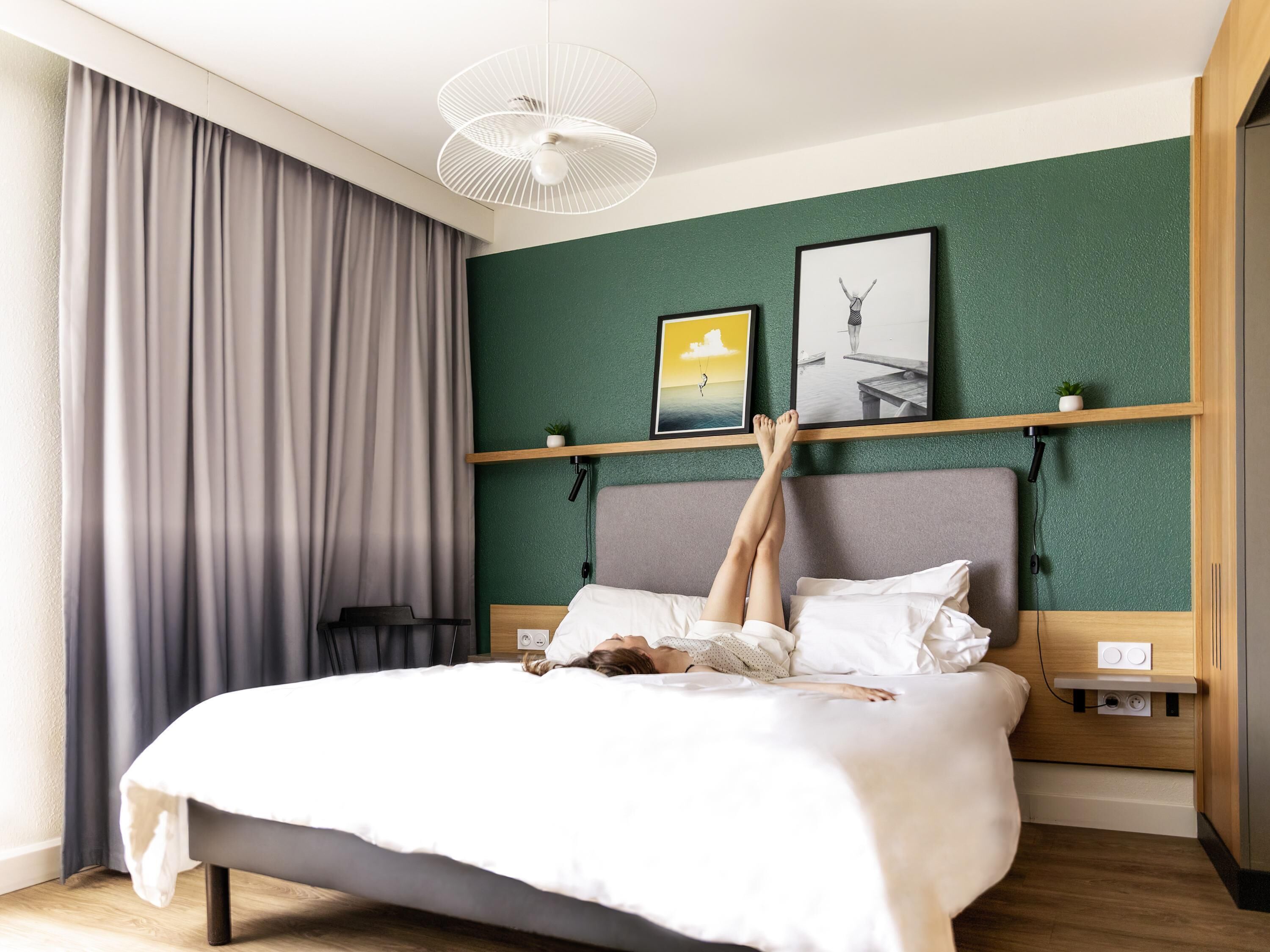 Een bed met een persoon erop, een plafondventilator, een muur met twee ingelijste foto's en een raam met gordijnen.