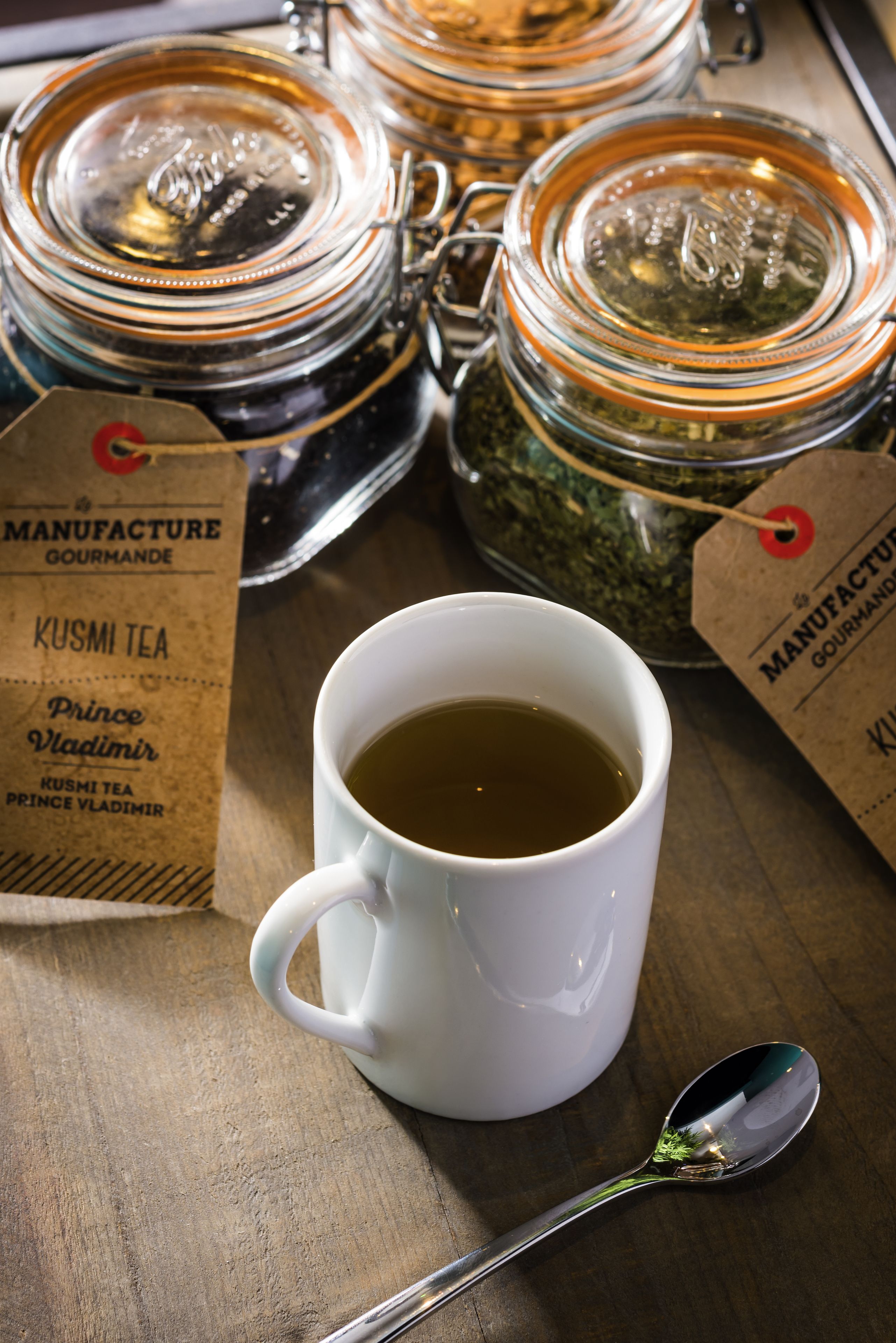 A white mug filled with tea, a spoon, and two glass jars with tea labels on a wooden surface.