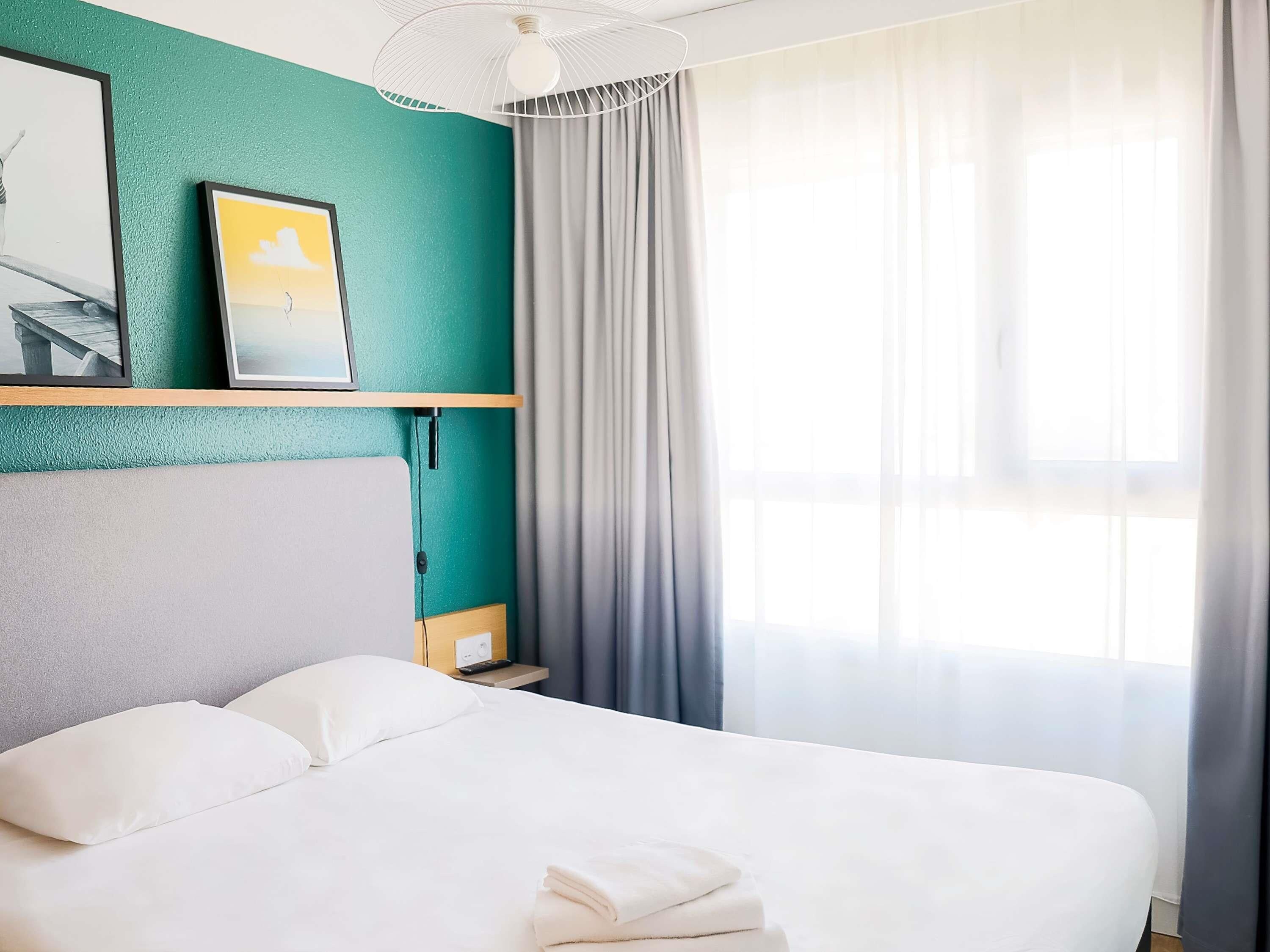 A hotel room with a bed, a nightstand, a wall-mounted shelf with pictures, a ceiling fan, and a window with curtains.