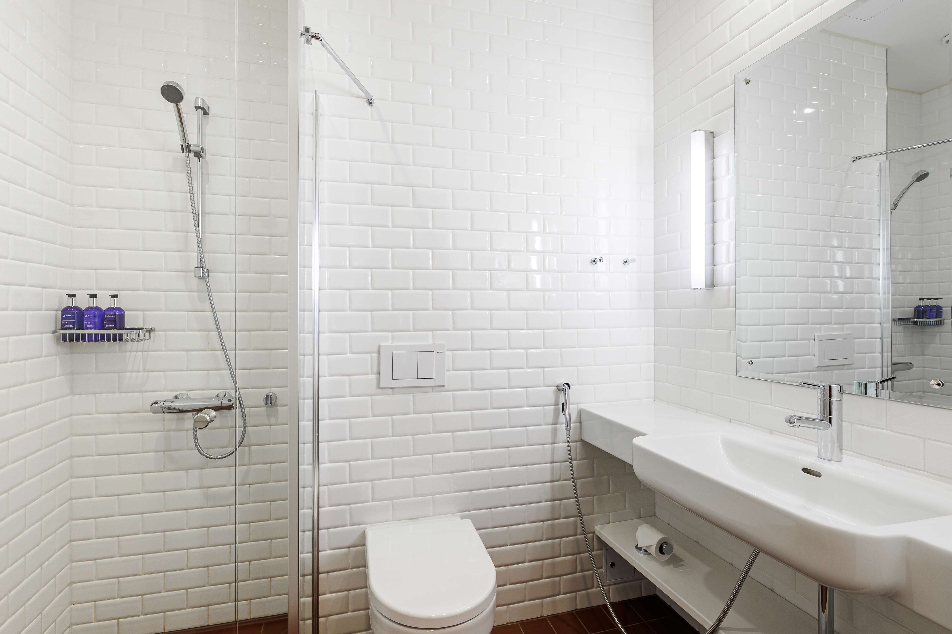 Un baño con azulejos blancos tipo metro, un inodoro blanco, una ducha con cerramiento de vidrio y un lavamanos blanco con espejo superior.