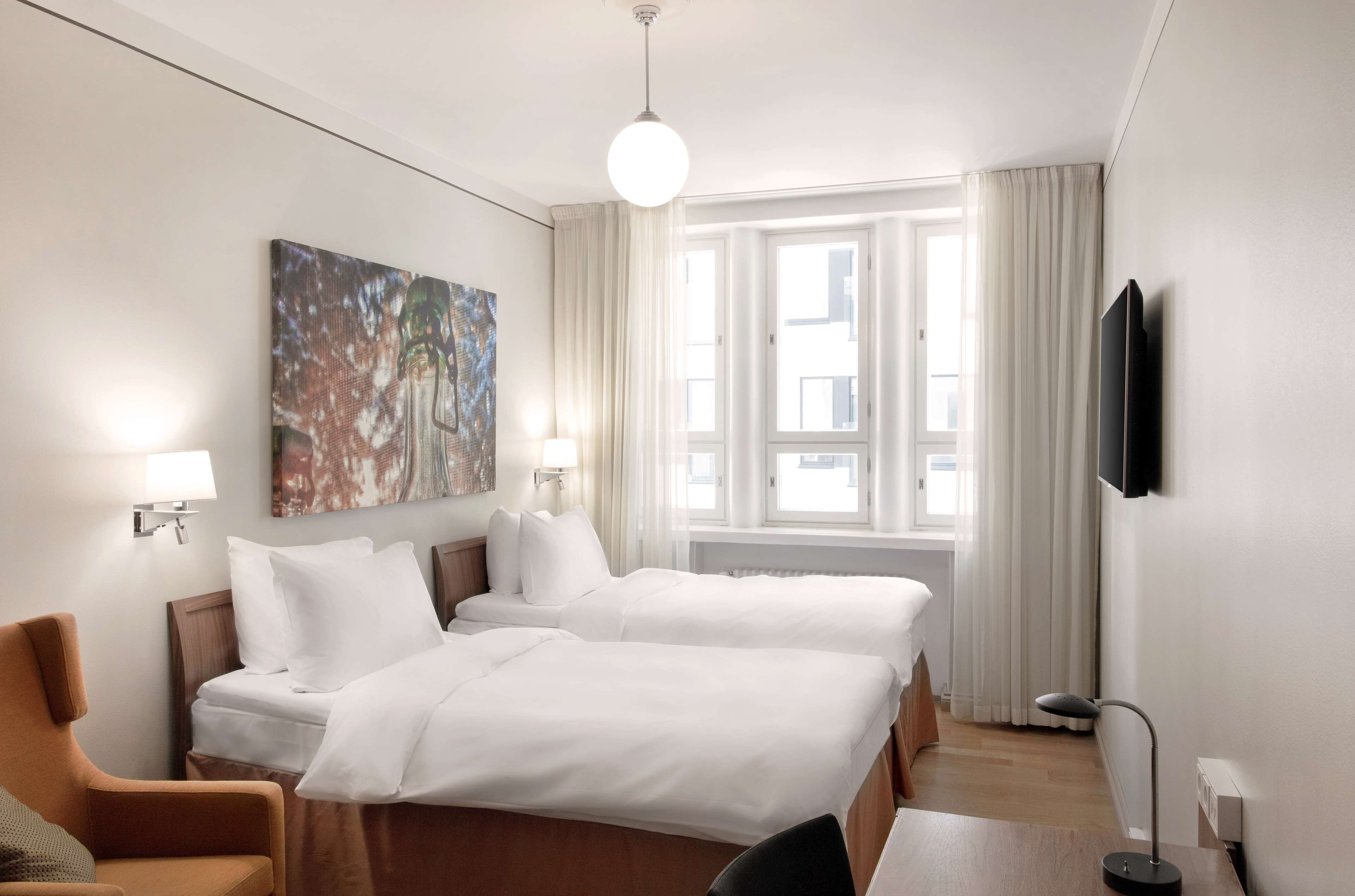Habitación de hotel con dos camas, un escritorio, una silla, una lámpara, un cuadro y un televisor en la pared.