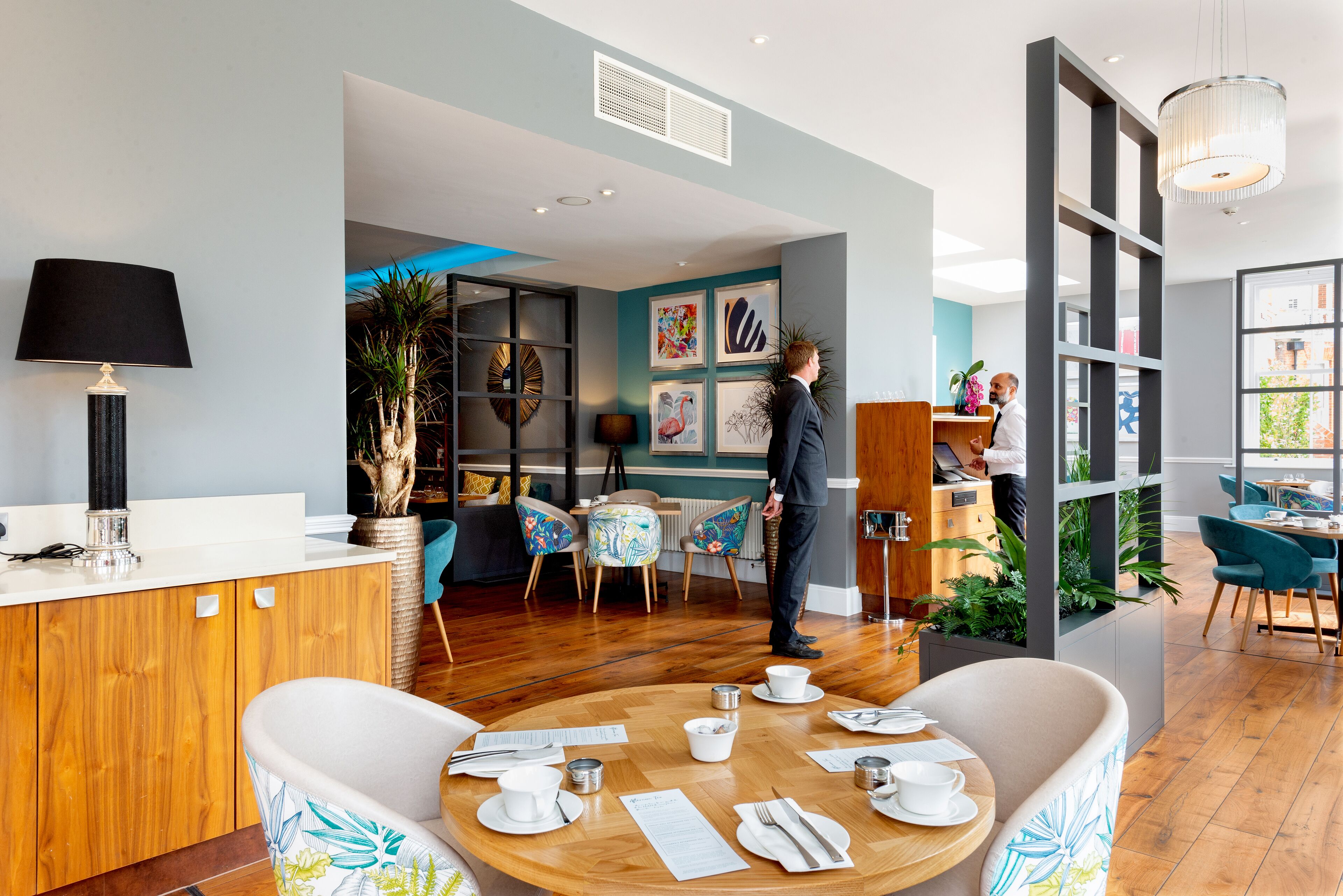 Un espace repas moderne avec une table ronde en bois dressée pour deux personnes, un coin bar avec un comptoir en bois, et une vue sur la cuisine grâce à une cloison en verre.