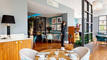 Un espace repas moderne avec une table ronde en bois dressée pour deux personnes, un coin bar avec un comptoir en bois, et une vue sur la cuisine grâce à une cloison en verre.