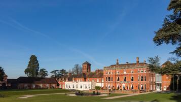 A large, historic red brick building with a central tower, surrounded by a well-maintained lawn and a small fountain.