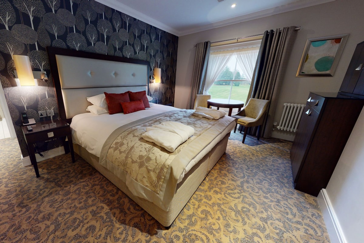 A hotel room with a large bed, a bedside table with a lamp, a chair, a small table, a radiator, and a window with curtains.