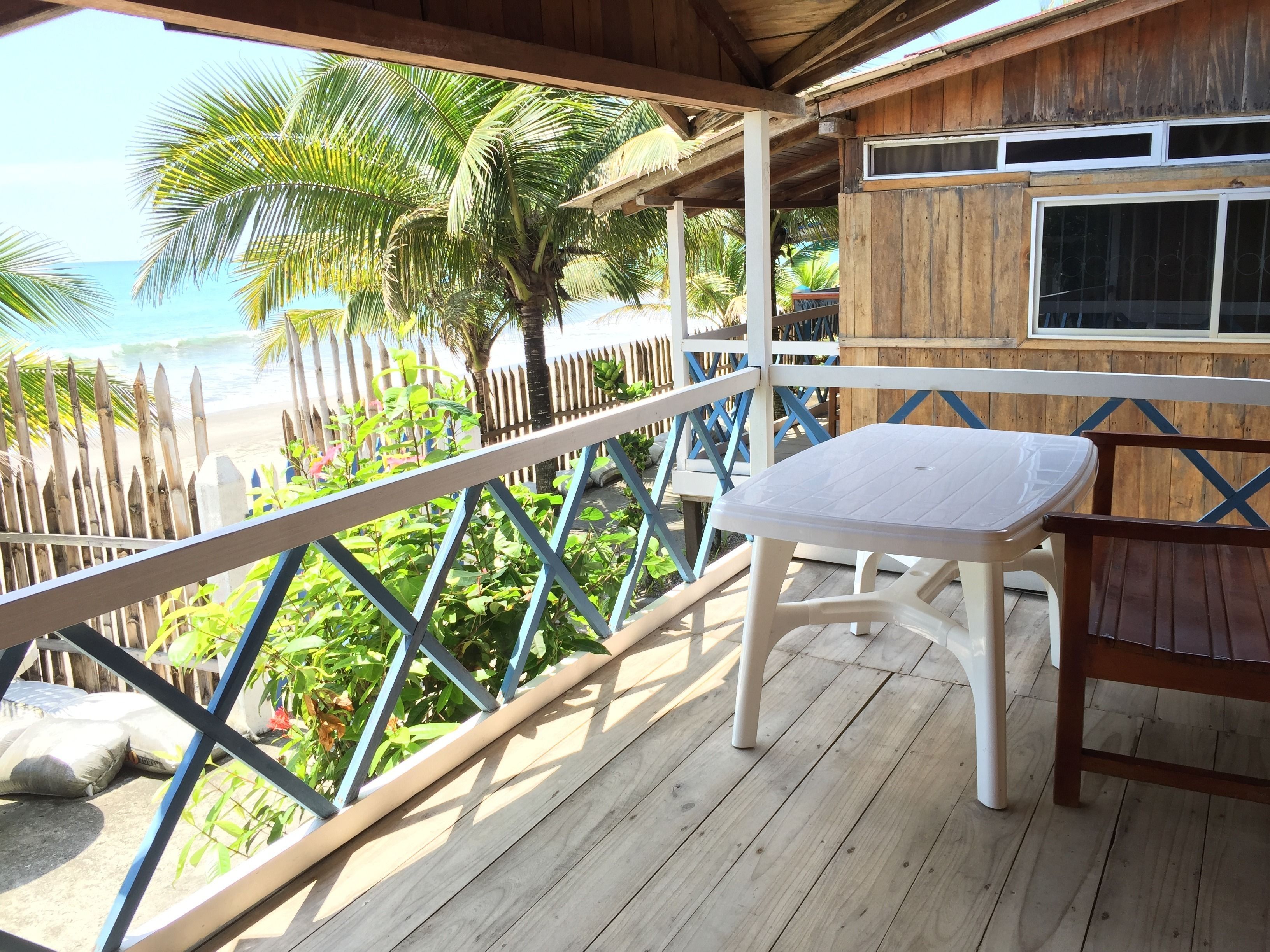 Una terraza de madera con una mesa y silla de plástico blanco, con vistas a una playa con palmeras y una valla de madera.