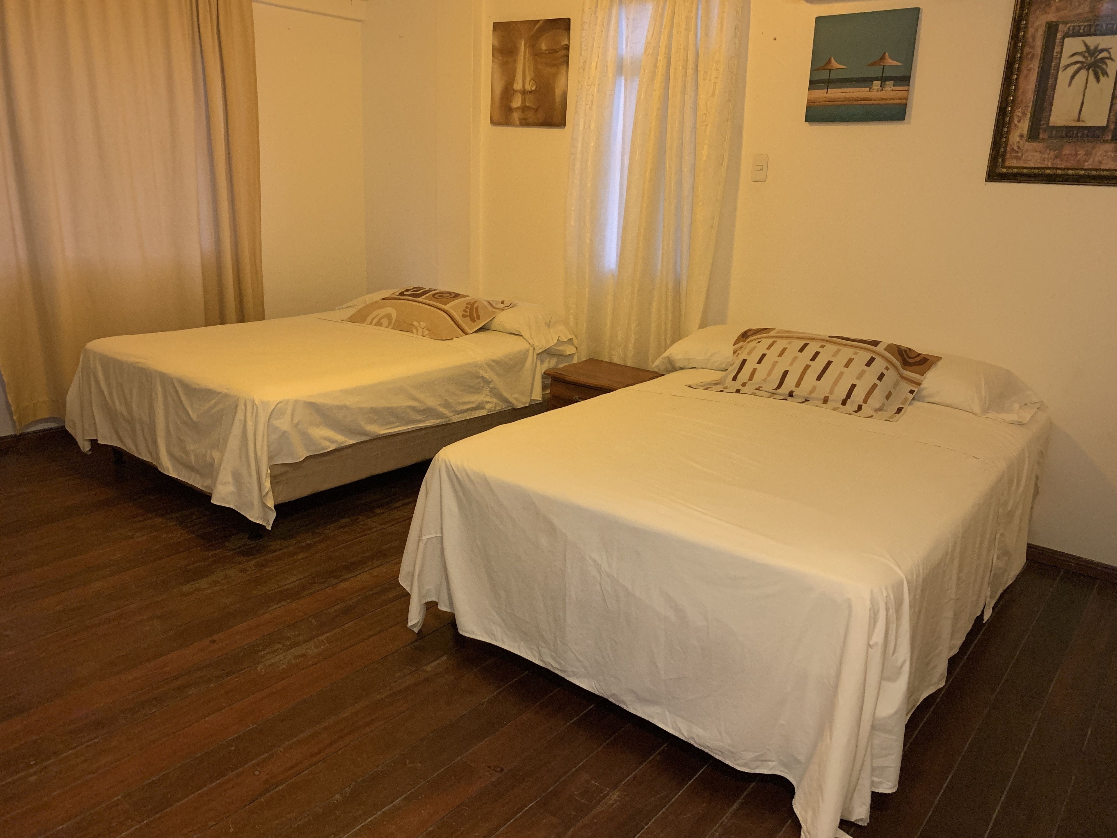 Habitación con dos camas individuales, piso de madera, ventana con cortinas y decoración de pared que incluye una escena de playa y una palmera.