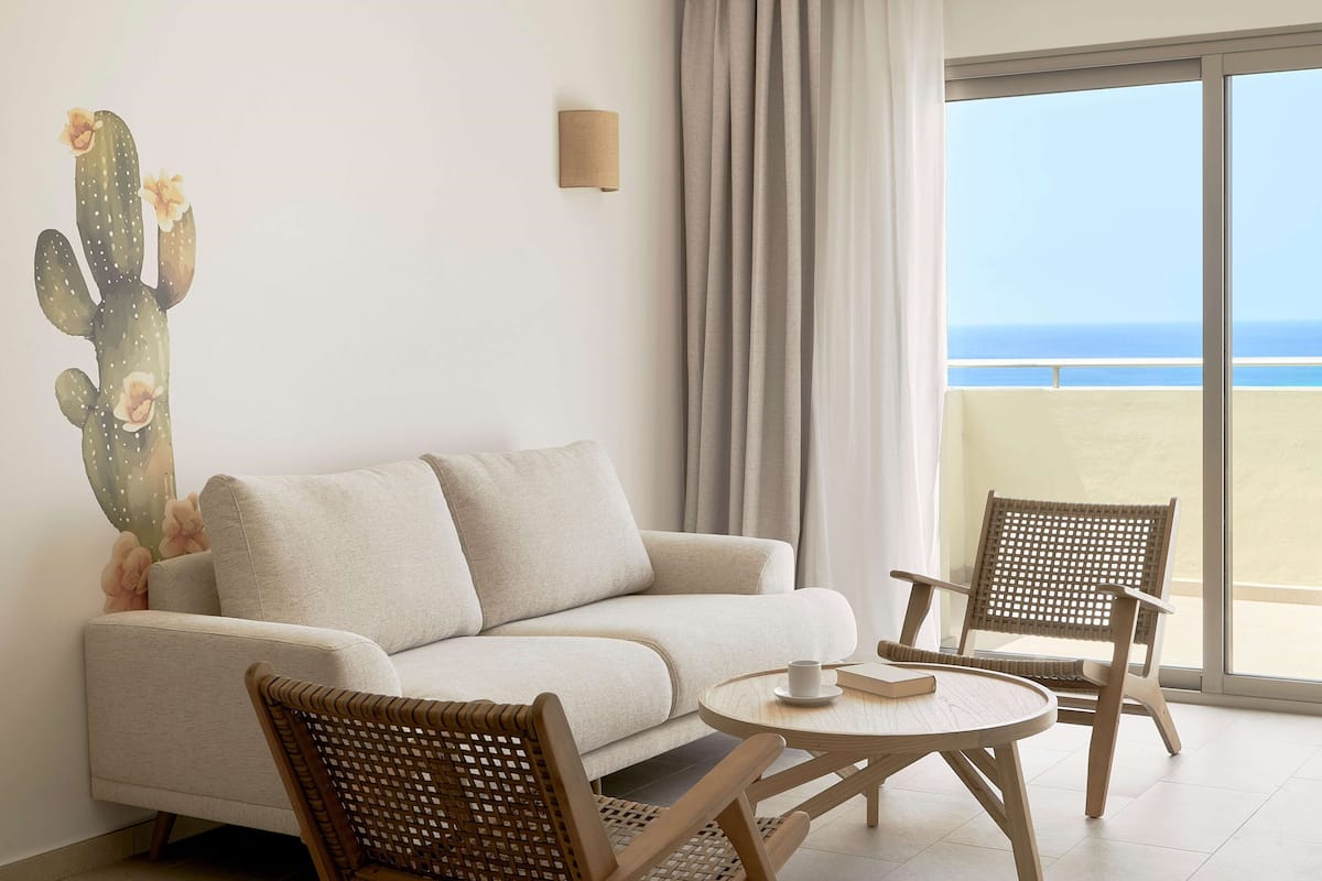 Une pièce comprenant un canapé, deux fauteuils en osier, une petite table, une œuvre d’art représentant un cactus et une vue sur la mer depuis un balcon.