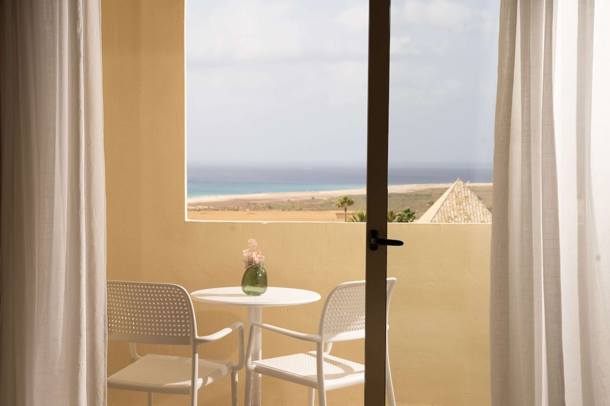 Un balcon avec deux chaises blanches et une petite table, donnant sur une plage et une structure pyramidale.