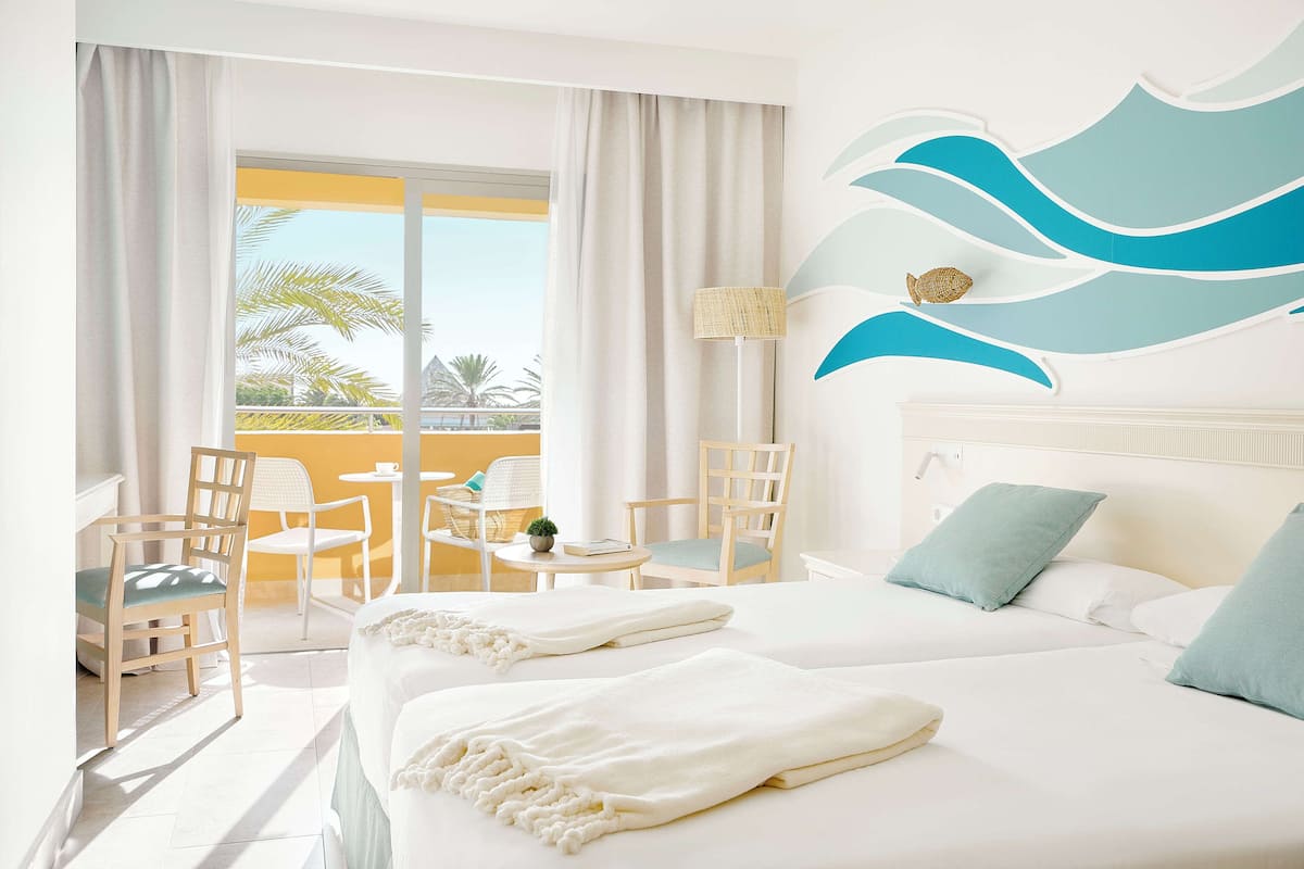 Une chambre d’hôtel avec un lit, deux chaises, une petite table, une lampe et une vue sur les palmiers, visible à travers de grandes fenêtres.