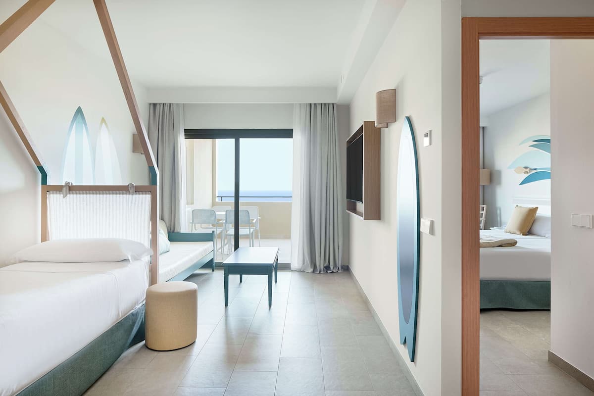 Une chambre d’hôtel avec un lit, une petite table, un banc, une télévision et une vue sur la mer.