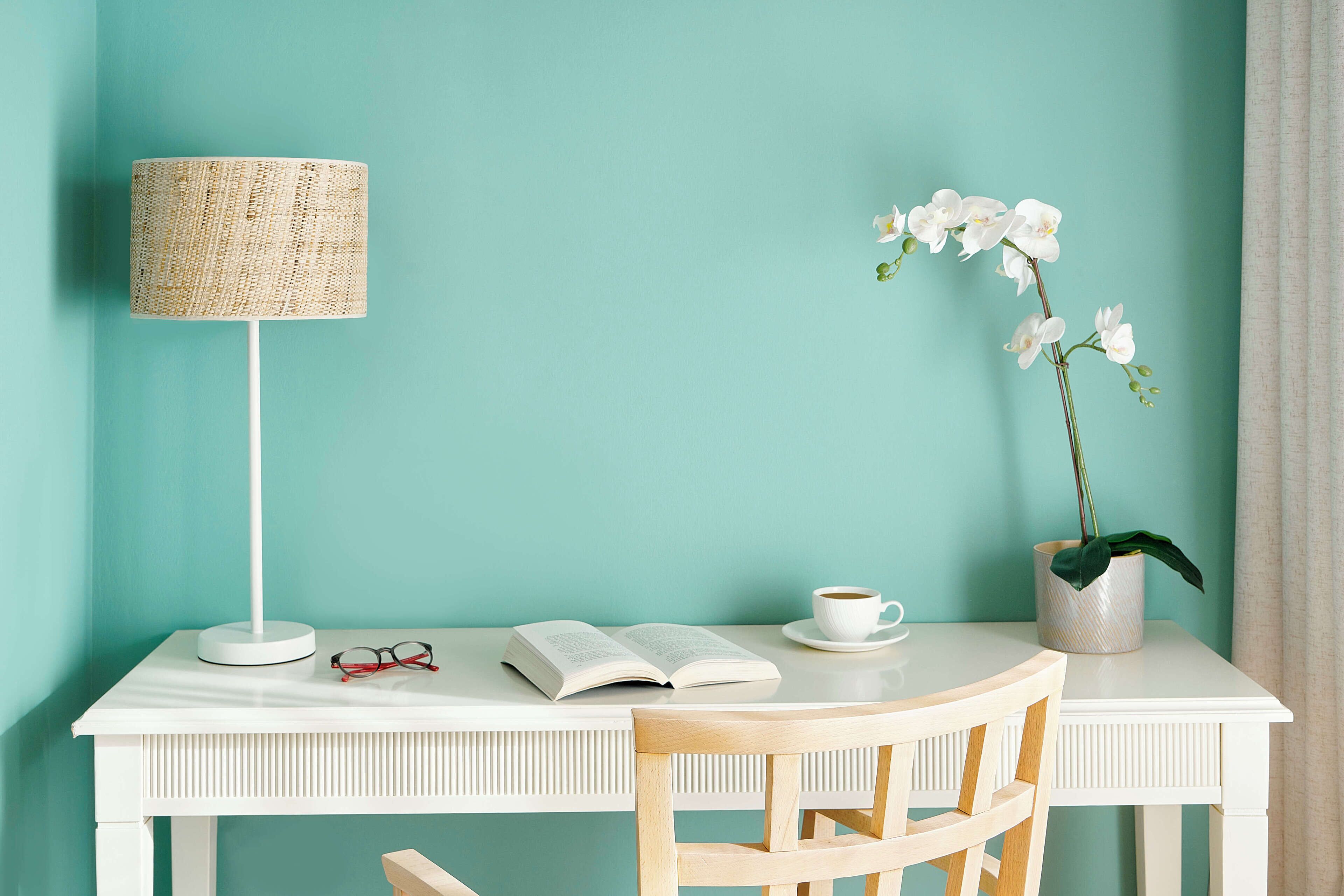 Un escritorio blanco con una lámpara, un libro abierto, un par de gafas, una taza y una orquídea en maceta.
