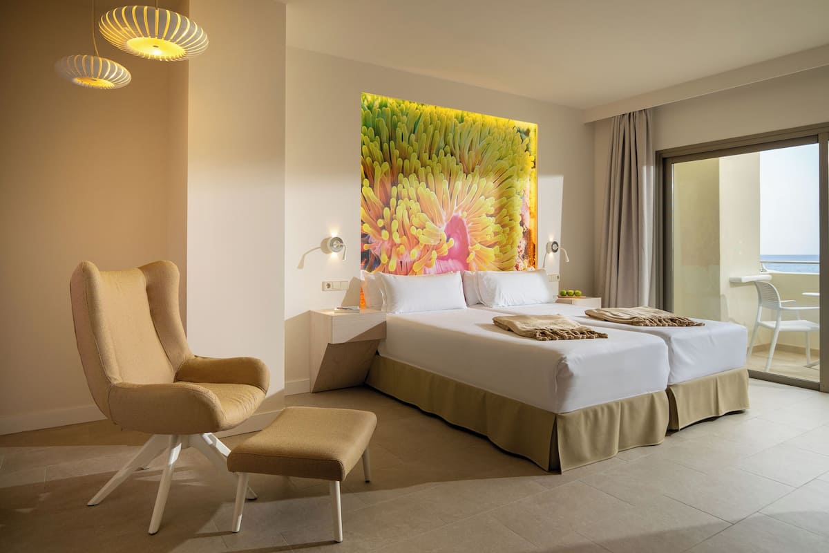 Une chambre d’hôtel moderne, dotée d’un grand lit, d’une chaise, d’une table de chevet et offrant une vue sur la mer depuis un balcon.