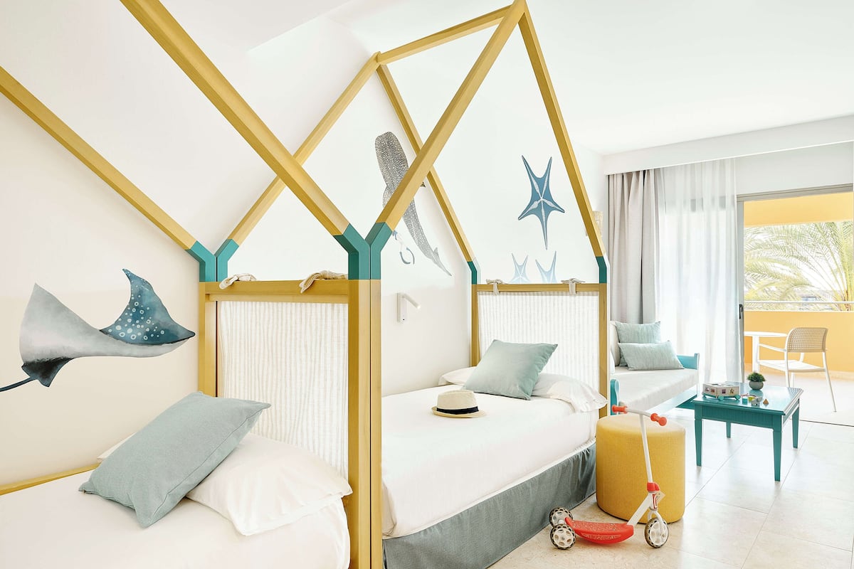 Une chambre à coucher avec un lit, des oreillers, un chapeau, une poussette et une fenêtre avec des rideaux.