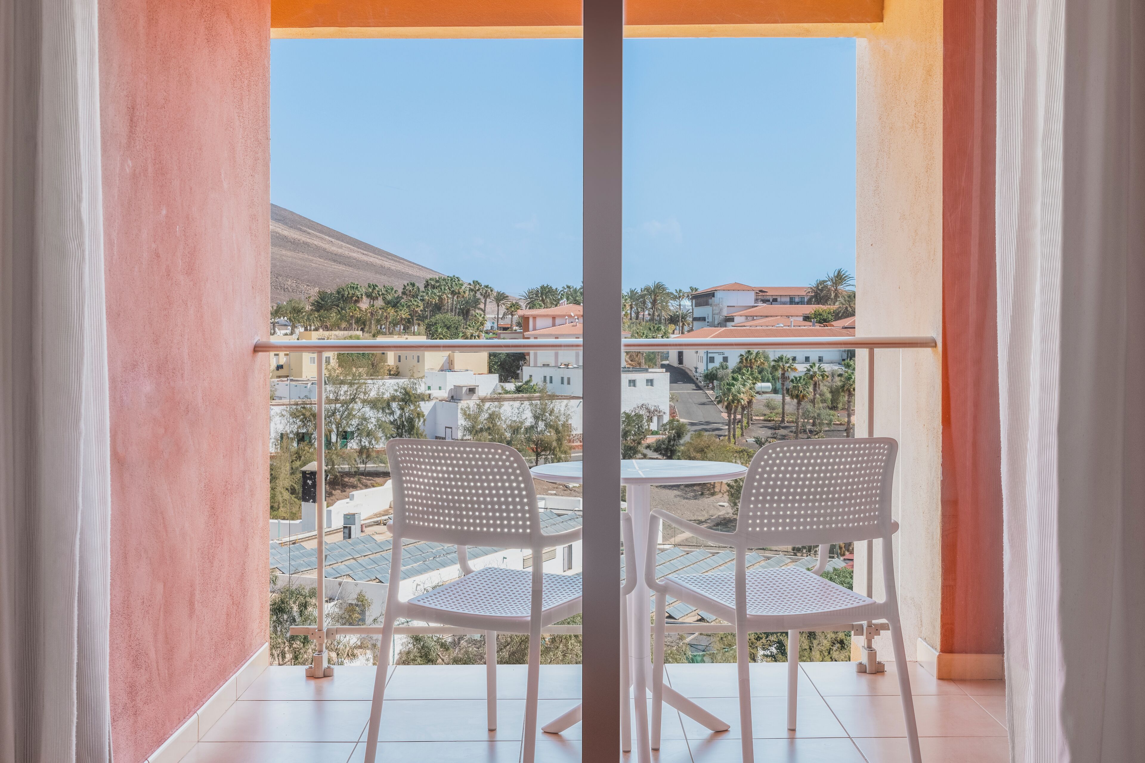 A balcony with two white chairs and a table, overlooking a residential area with houses and a mountain.