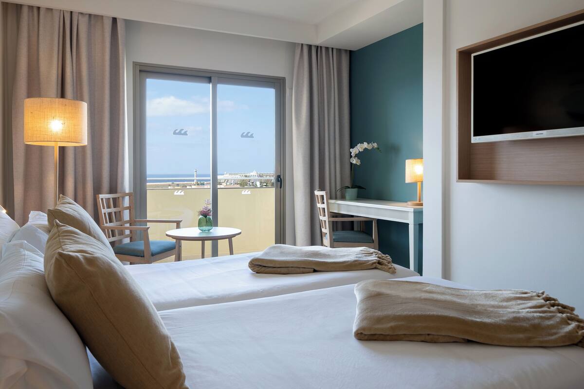 Une chambre d’hôtel avec un lit, un bureau, une chaise, une lampe et une vue sur la plage.