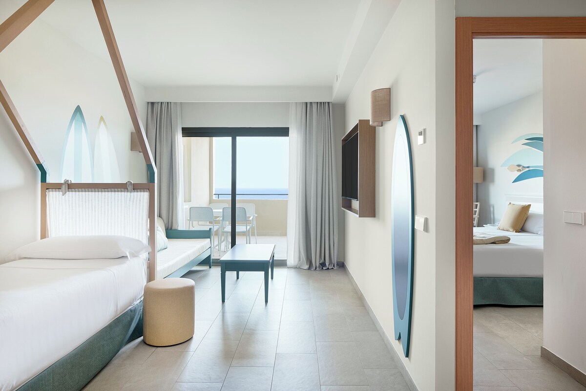 A hotel room with a bed, a small table, a bench, a TV, and a view of the sea.