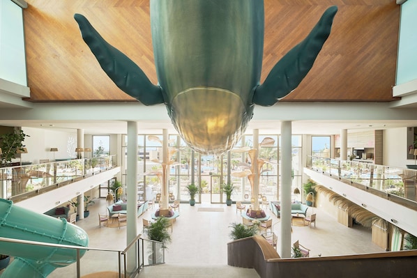 A spacious atrium with a central glass ceiling, multiple levels with seating areas, and a visible pool area. - Iberostar Waves Gaviotas Park -All Inclusive (Pajara)