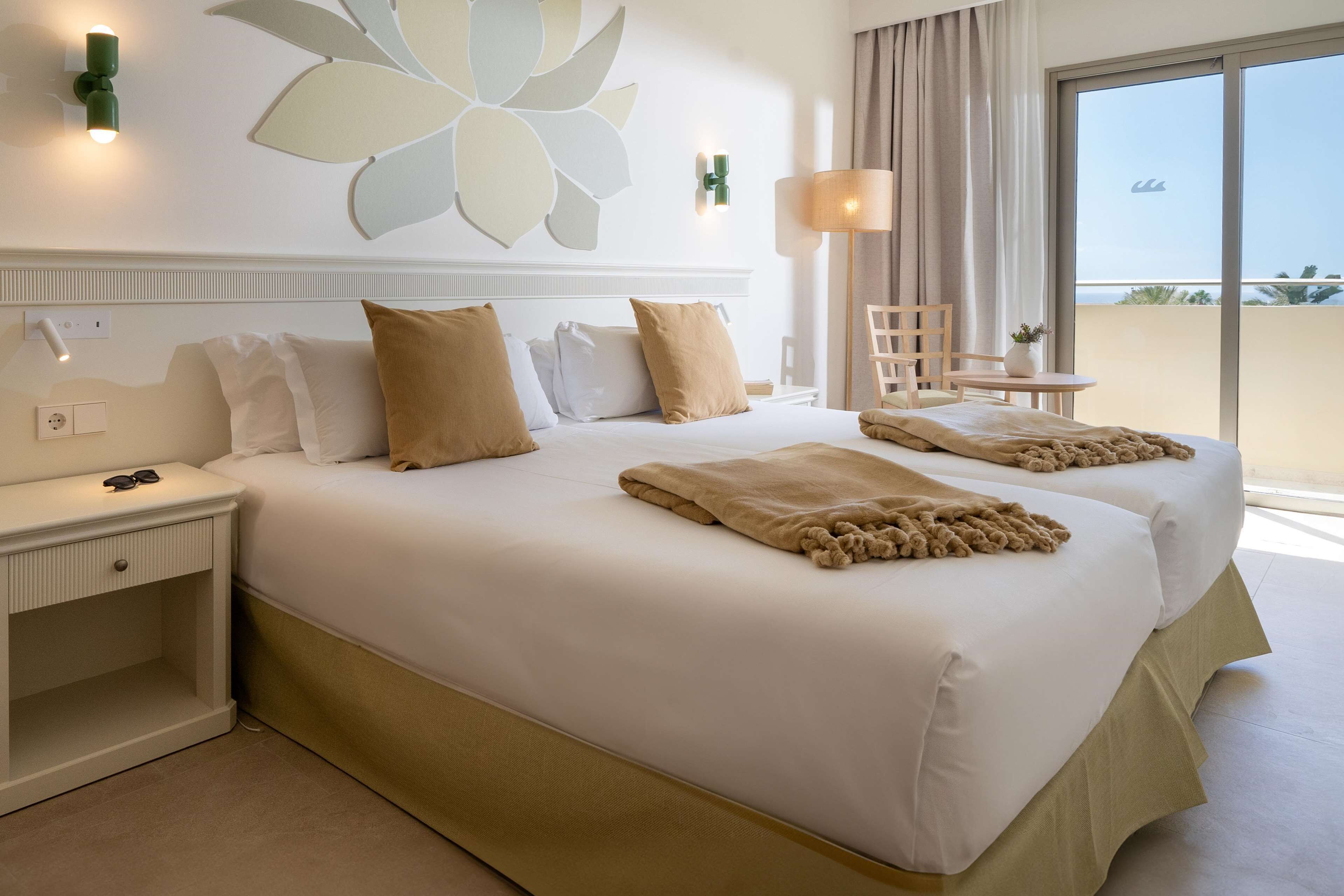 Habitación de hotel con una cama grande, una mesita de noche, una silla, una pequeña mesa con una planta y vistas al exterior.