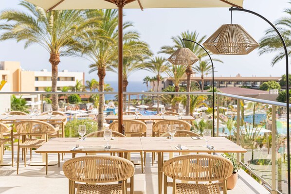 A terrace with tables and chairs, overlooking a pool area with palm trees and buildings in the background. - Iberostar Waves Gaviotas Park -All Inclusive (Pajara)