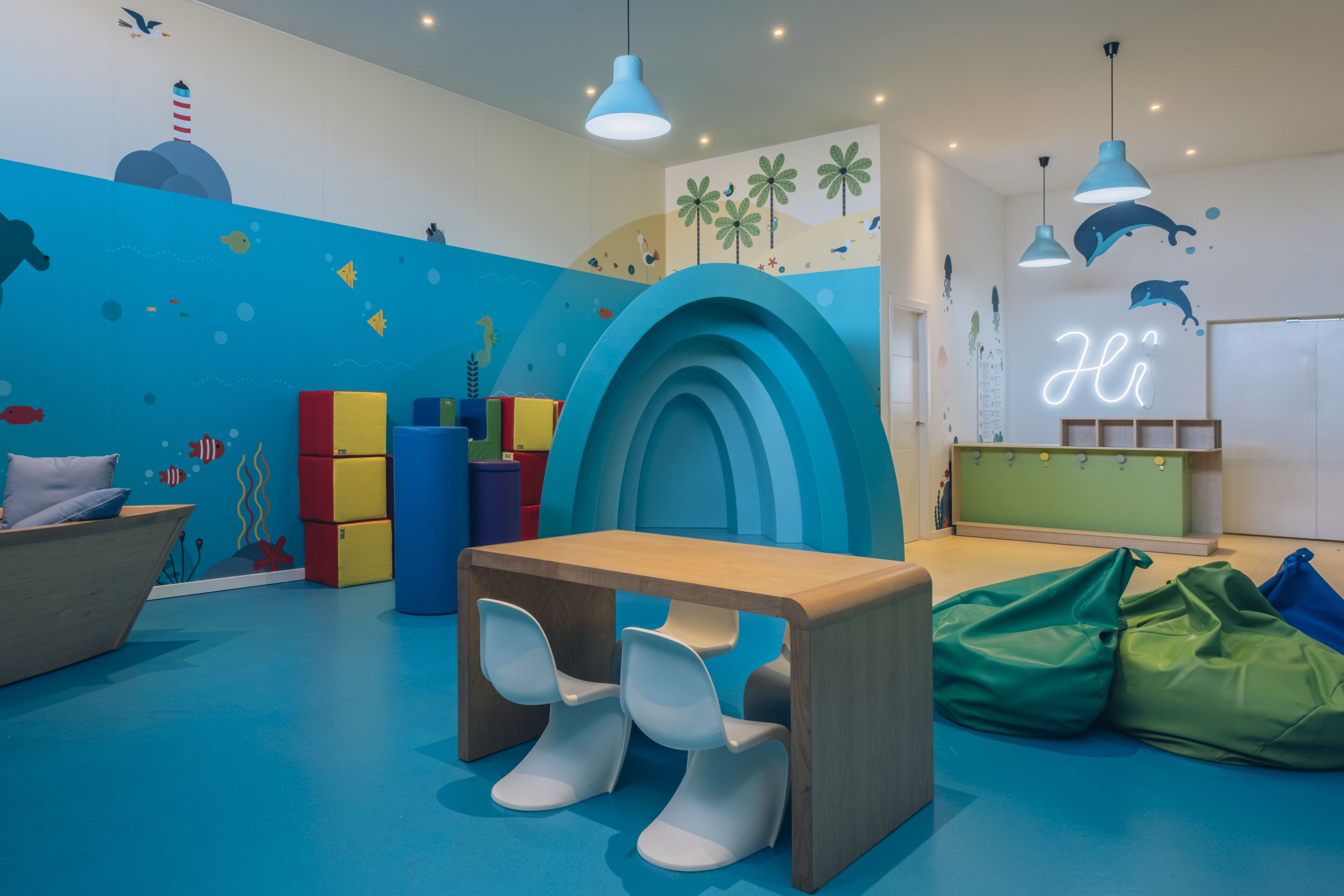 Sala de juegos infantil con pared azul, estructuras de juego coloridas, una mesa de madera y un letrero de neón que dice 'Hi'.
