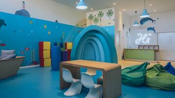 Sala de juegos infantil con pared azul, estructuras de juego coloridas, una mesa de madera y un letrero de neón que dice 'Hi'.