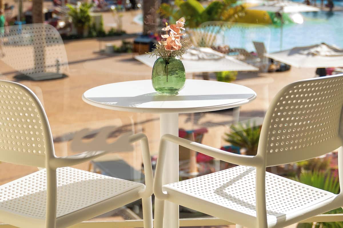 Une table ronde ornée d’un vase de fleurs, deux chaises blanches et une vue sur une piscine et des palmiers.