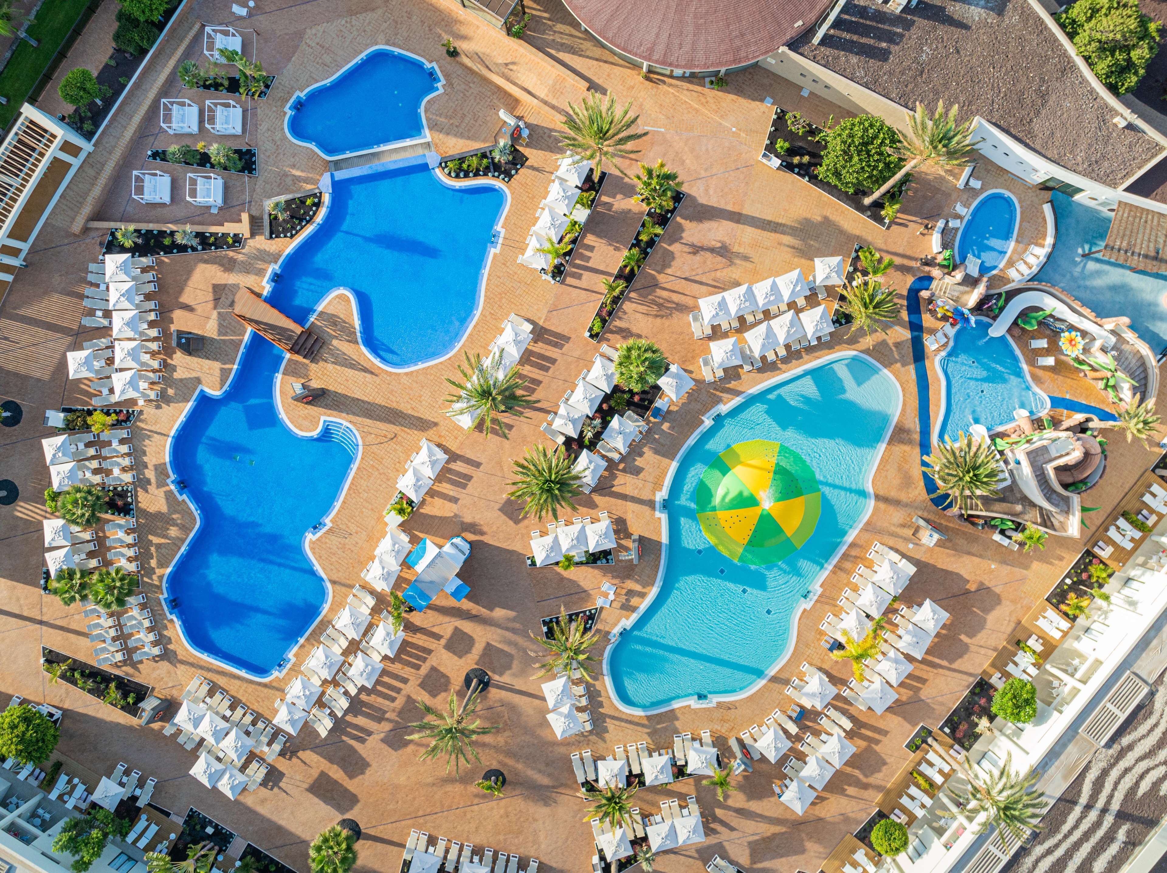 Vista aérea de un complejo turístico con múltiples piscinas, tumbonas y palmeras.
