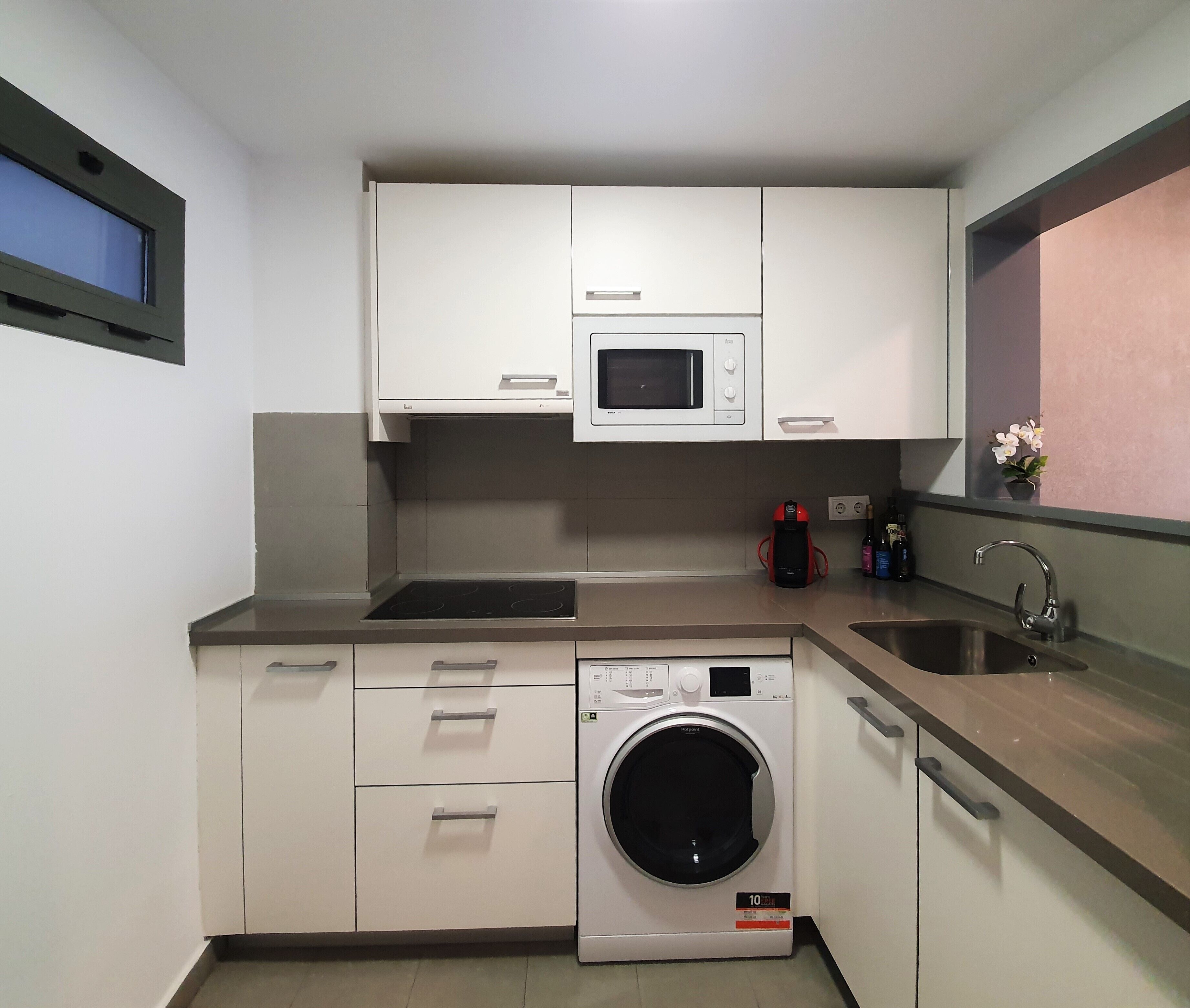 Cocina compacta con armarios blancos, microondas, lavadora integrada, pileta y ventana con vistas.
