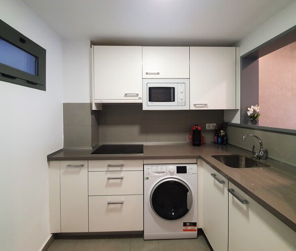 A compact kitchen with white cabinets, a microwave, a built-in washing machine, a sink, and a window with a view. - Iberflat Vega De Triana (Camas)